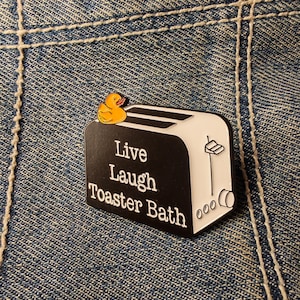 Könnte beinhalten: Schwarz-weißer Emaille-Pin in Form eines Toasters mit einer gelben Quietscheente oben. Der Pin trägt die Worte "Live Laugh Toaster Bath" in weißer Schrift.