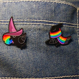 May include: Two enamel pins depicting black birds. One bird wears a purple witch's hat and has a rainbow beak. The other bird wears a rainbow scarf. Both pins are set against a blue denim background.
