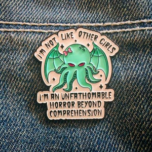May include: A metal enamel pin featuring a light green octopus-like creature with bat wings, a small pink bow, and red eyes. The text reads: I'M NOT LIKE OTHER GIRLS I'M AN UNFATHOMABLE HORROR BEYOND COMPREHENSION. The background is a pale cream colour.