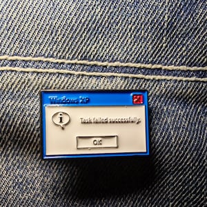 Może przedstawiać: Niebiesko-biała emaliowana przypinka w kształcie komunikatu o błędzie systemu Windows XP. Przypinka zawiera tekst "Windows XP", "Task failed successfully" i przycisk "OK". Przypinka jest umieszczona na tle dżinsu.