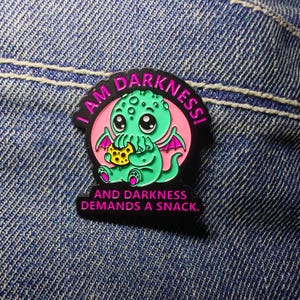 Puede incluir: Pin de esmalte negro con un monstruo de dibujos animados verde, ojos grandes y alas rosas, sosteniendo una galleta. El pin tiene el texto "I AM DARKNESS!" y "AND DARKNESS DEMANDS A SNACK."