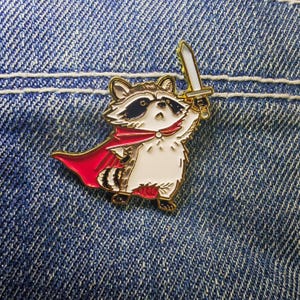 May include: A gold-coloured metal pin depicting a cartoon raccoon wearing a red cape and holding a sword. The raccoon has black markings around its eyes and a white body. The pin is set against a blue denim background.