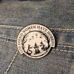 May include: A circular enamel pin in black and white, featuring the text "GOD FORBID WOMEN HAVE HOBBIES" around a design of three witches in a forest. The pin is displayed on a denim fabric.