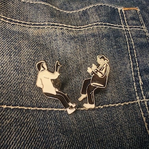 May include: Two enamel pins featuring figures in black and white. One figure wears a white jacket and black trousers, the other a black jacket and black trousers. The pins are set against a blue denim background.
