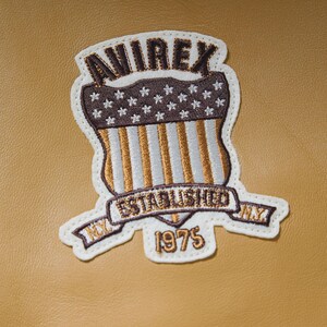 May include: Embroidered patch with the text "AVIREX ESTABLISHED NY 1975". The patch features a shield design with a brown and white color scheme, including stars and stripes, on a tan background.