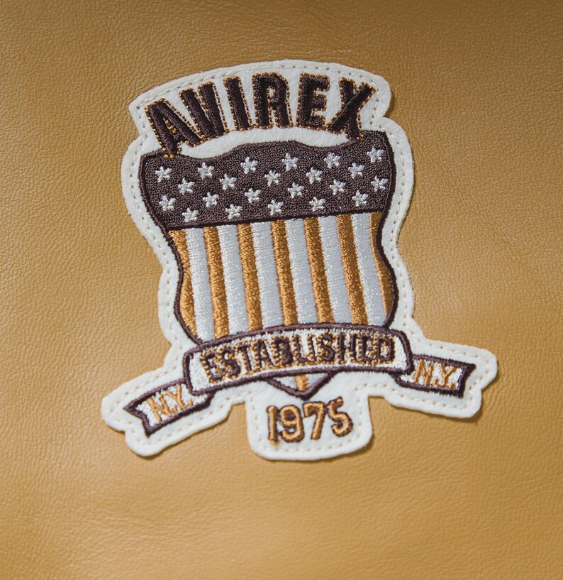 May include: Embroidered patch with the text "AVIREX ESTABLISHED NY 1975". The patch features a shield design with a brown and white color scheme, including stars and stripes, on a tan background.