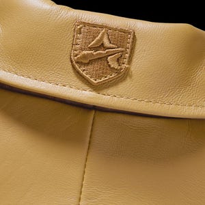 May include: Close-up of a tan leather item with a stitched emblem. The emblem is a shield shape with a detailed embroidered design in a matching tan color. The leather has a subtle texture.
