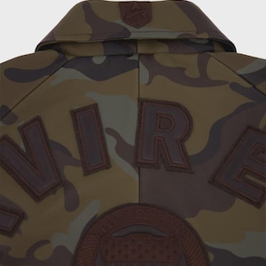 May include: A camouflage jacket featuring a brown leather collar with a small embossed logo. The back displays large, dark brown leather letters spelling out part of the word "WIRE" and a circular leather patch with an embossed shield design.