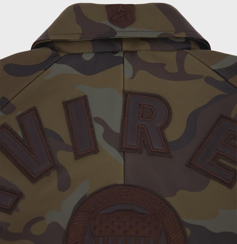 May include: A camouflage jacket featuring a brown leather collar with a small embossed logo. The back displays large, dark brown leather letters spelling out part of the word "WIRE" and a circular leather patch with an embossed shield design.
