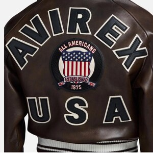 May include: A dark brown leather jacket featuring the text AVIREX in black letters across the top and USA in black letters across the bottom. A central circular patch displays a shield with stars and stripes, the text ALL AMERICANS, ESTABLISHED 1975, and N.Y.
