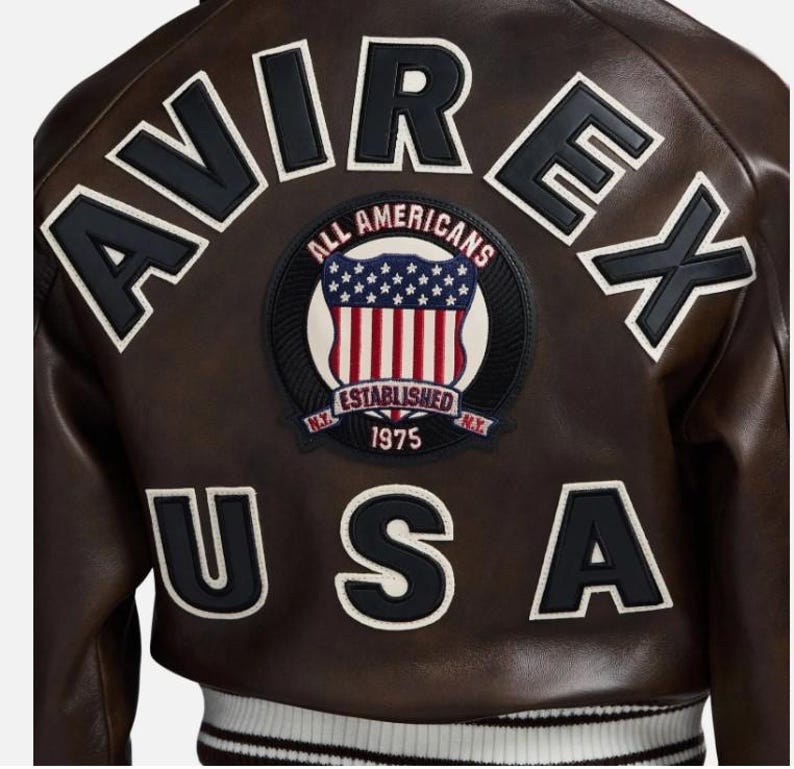 May include: A dark brown leather jacket featuring the text AVIREX in black letters across the top and USA in black letters across the bottom. A central circular patch displays a shield with stars and stripes, the text ALL AMERICANS, ESTABLISHED 1975, and N.Y.
