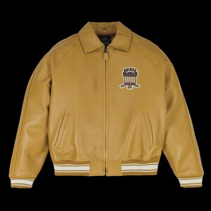 May include: Mustard-colored leather jacket with a zip-up front, collared neckline, and ribbed cuffs and hem with white stripes. Features a patch on the chest with the text "AVIREX" and a patriotic design. A classic bomber jacket style.