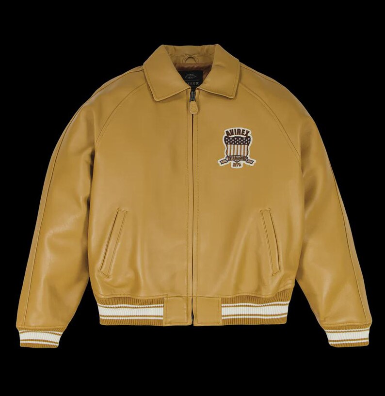 May include: Mustard-colored leather jacket with a zip-up front, collared neckline, and ribbed cuffs and hem with white stripes. Features a patch on the chest with the text "AVIREX" and a patriotic design. A classic bomber jacket style.