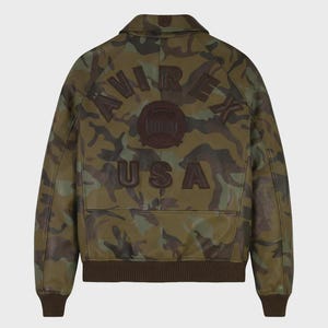 May include: A camouflage print leather jacket featuring a brown collar, cuffs, and hem. The back displays dark brown embossed lettering that reads AVIREX USA, along with a circular emblem in the center, all set against a green and brown pattern.