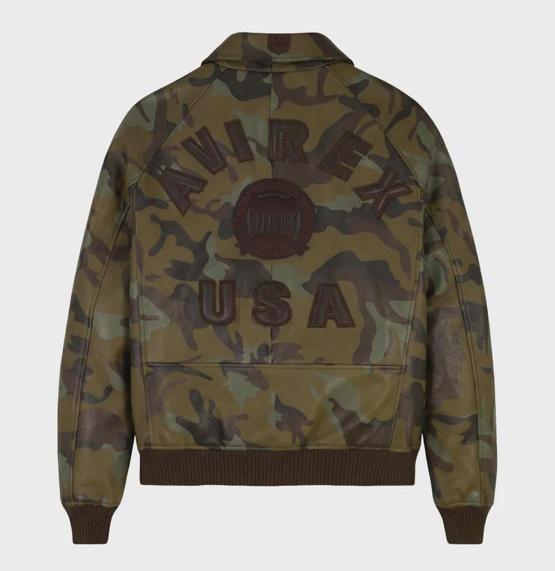 May include: A camouflage print leather jacket featuring a brown collar, cuffs, and hem. The back displays dark brown embossed lettering that reads AVIREX USA, along with a circular emblem in the center, all set against a green and brown pattern.