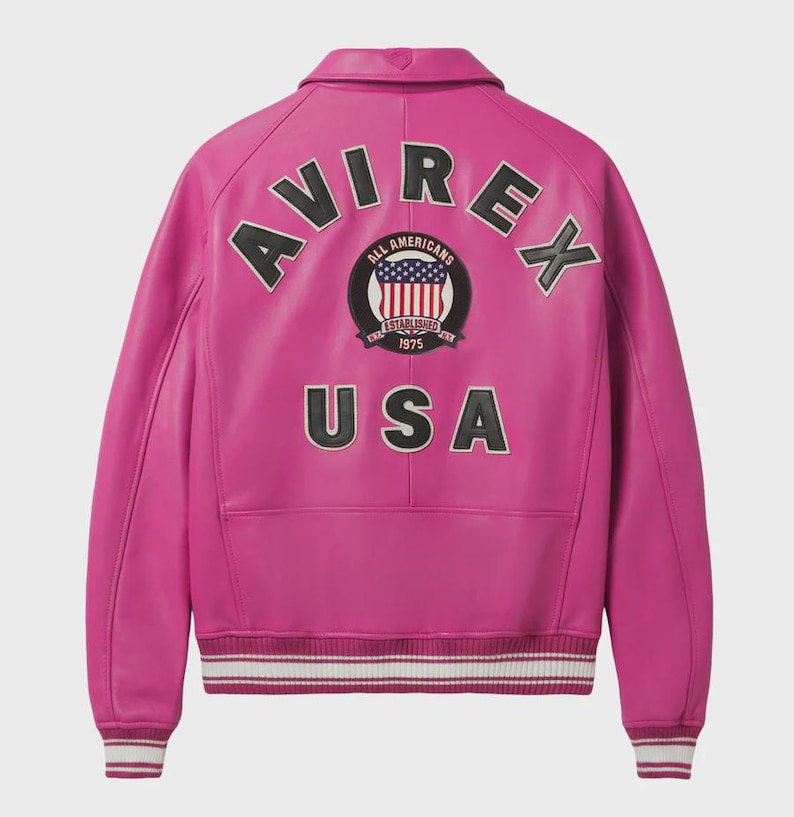 Op de afbeelding: Een felroze leren jas met het woord "AVIREX" over de schouders. De achterkant heeft een cirkelvormige patch met de Amerikaanse vlag en de tekst "ALL AMERICANS ESTABLISHED 1975 USA". De jas heeft een geribbelde zoom en manchetten.