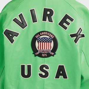 K&ouml;nnte beinhalten: Eine leuchtend gr&uuml;ne Jacke mit schwarz-wei&szlig;en Buchstaben und Aufn&auml;hern. Das Wort "AVIREX" steht oben und "USA" unten. Ein kreisf&ouml;rmiger Aufn&auml;her in der Mitte zeigt die amerikanische Flagge und die Worte "ALL AMERICANS ESTABLISHED 1975 N.Y."
