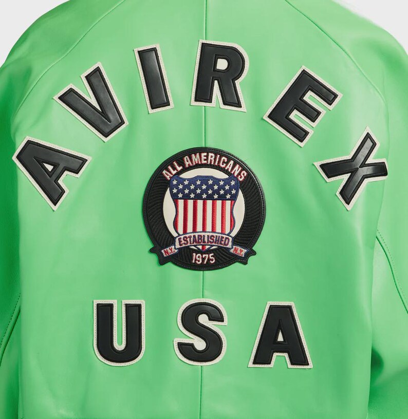 K&ouml;nnte beinhalten: Eine leuchtend gr&uuml;ne Jacke mit schwarz-wei&szlig;en Buchstaben und Aufn&auml;hern. Das Wort "AVIREX" steht oben und "USA" unten. Ein kreisf&ouml;rmiger Aufn&auml;her in der Mitte zeigt die amerikanische Flagge und die Worte "ALL AMERICANS ESTABLISHED 1975 N.Y."