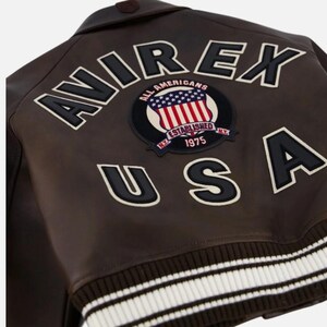 May include: A dark brown leather jacket featuring the text AVIREX USA in large black letters with white trim. A circular patch in the center displays the text ALL AMERICANS, ESTABLISHED 1975, and N.Y. with a graphic of the American flag. The hem is ribbed.