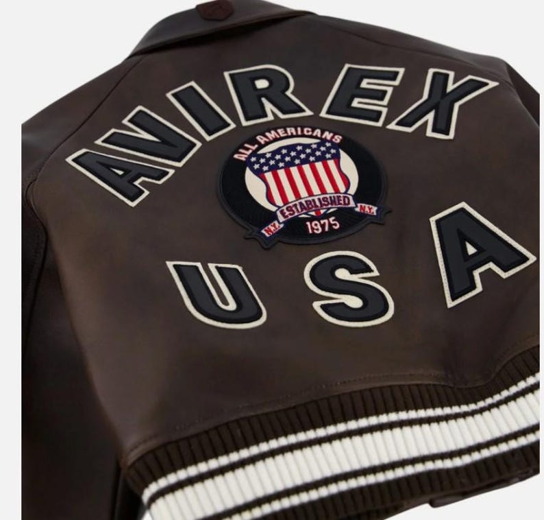 May include: A dark brown leather jacket featuring the text AVIREX USA in large black letters with white trim. A circular patch in the center displays the text ALL AMERICANS, ESTABLISHED 1975, and N.Y. with a graphic of the American flag. The hem is ribbed.