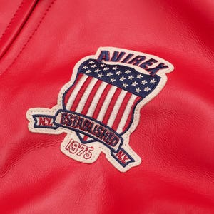 May include: Close-up of a red leather jacket with a stitched patch. The patch features a shield design with the American flag, the word "AVIREX" and the text "ESTABLISHED 1975 NY". The jacket has a textured surface.