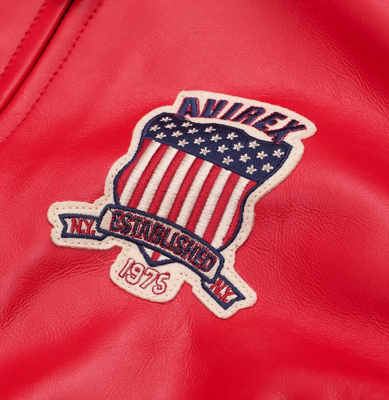 May include: Close-up of a red leather jacket with a stitched patch. The patch features a shield design with the American flag, the word "AVIREX" and the text "ESTABLISHED 1975 NY". The jacket has a textured surface.
