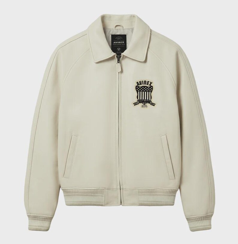 Pode incluir: Casaco de couro creme com gola e fecho de correr frontal. Apresenta um emblema a preto e branco no peito com o texto "AVIREX" e "1975". Inclui dois bolsos frontais e punhos e bainha canelados.