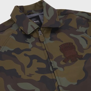 May include: A camouflage print jacket featuring a pointed collar, a full-length front zipper with a branded pull tab, and a dark brown embroidered patch on the chest. The interior label displays the text Authentic 1975 Avirex.