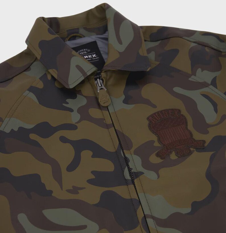 May include: A camouflage print jacket featuring a pointed collar, a full-length front zipper with a branded pull tab, and a dark brown embroidered patch on the chest. The interior label displays the text Authentic 1975 Avirex.