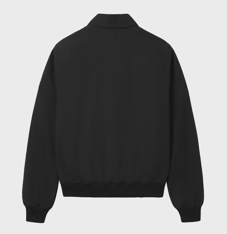 May include: A solid black jacket featuring a pointed collar, long sleeves with ribbed cuffs, and a ribbed hem. The garment has a clean, minimalist design with a small, subtle tonal logo embroidered on the center back of the collar area.