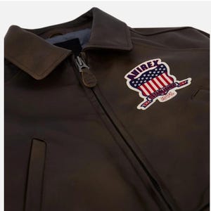 May include: A dark brown leather jacket with a full-length front zipper and a pointed collar. A patch on the chest features the text AVIREX ESTABLISHED 1975 with a graphic of the American flag. The zipper pull has a small leather tab attached.