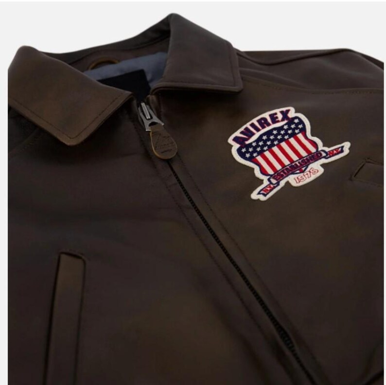 May include: A dark brown leather jacket with a full-length front zipper and a pointed collar. A patch on the chest features the text AVIREX ESTABLISHED 1975 with a graphic of the American flag. The zipper pull has a small leather tab attached.