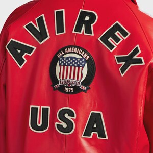 May include: Red leather jacket with the word "AVIREX" across the top and "USA" at the bottom in black letters with white trim. A circular patch in the center reads "ALL AMERICANS ESTABLISHED 1975" with a design of the American flag.