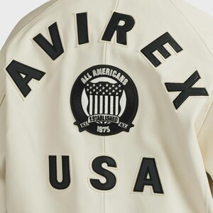 Pode incluir: Casaco creme com letras pretas que soletram "AVIREX" e "USA". Um patch circular apresenta o texto "ALL AMERICANS ESTABLISHED 1975 N.Y." com um design de estrelas e listras. O casaco tem um estilo universit&aacute;rio cl&aacute;ssico.