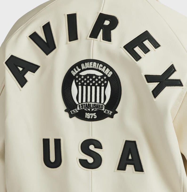 Pode incluir: Casaco creme com letras pretas que soletram "AVIREX" e "USA". Um patch circular apresenta o texto "ALL AMERICANS ESTABLISHED 1975 N.Y." com um design de estrelas e listras. O casaco tem um estilo universit&aacute;rio cl&aacute;ssico.