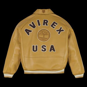 May include: Mustard-colored leather jacket with the word "AVIREX" arched across the back in brown lettering, above a circular emblem and the word "USA". The jacket has a collar, ribbed cuffs, and a white and brown striped hem.