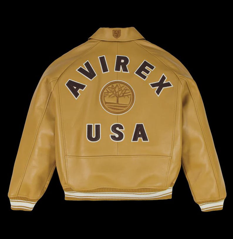 May include: Mustard-colored leather jacket with the word "AVIREX" arched across the back in brown lettering, above a circular emblem and the word "USA". The jacket has a collar, ribbed cuffs, and a white and brown striped hem.