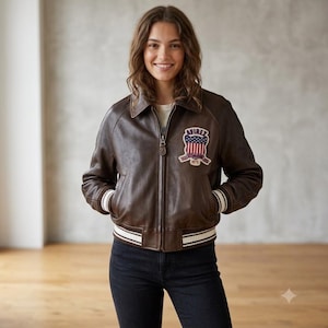 May include: A dark brown leather bomber jacket featuring a front zipper closure, a classic collar, and ribbed cuffs and hem with white stripes. An embroidered patch on the chest displays the text AVIREX with a shield and American flag design.