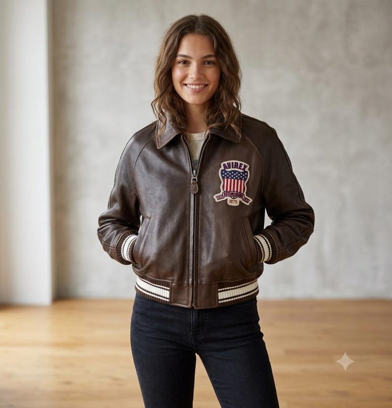 May include: A dark brown leather bomber jacket featuring a front zipper closure, a classic collar, and ribbed cuffs and hem with white stripes. An embroidered patch on the chest displays the text AVIREX with a shield and American flag design.
