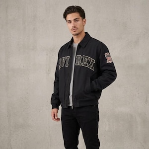 May include: A black zip-up jacket with a collar, featuring the text AVI REX in large, arched, light-colored block letters across the chest and a small, rectangular patch with a flag graphic on the upper left sleeve, worn over a gray shirt.