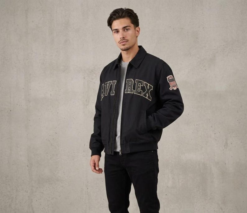 May include: A black zip-up jacket with a collar, featuring the text AVI REX in large, arched, light-colored block letters across the chest and a small, rectangular patch with a flag graphic on the upper left sleeve, worn over a gray shirt.