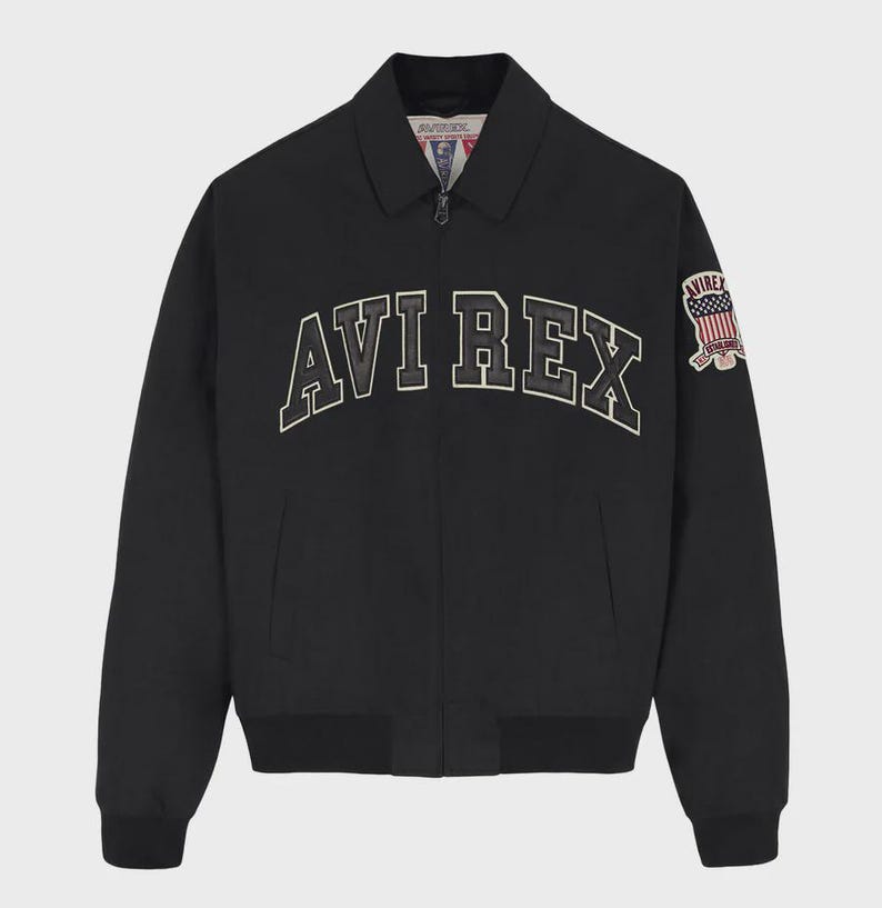 May include: A black zip-up jacket featuring a classic collar and ribbed cuffs. The chest displays the text AVI REX in large, arched, white-outlined black lettering. A patch on the left sleeve features an American flag and the text AVIREX.