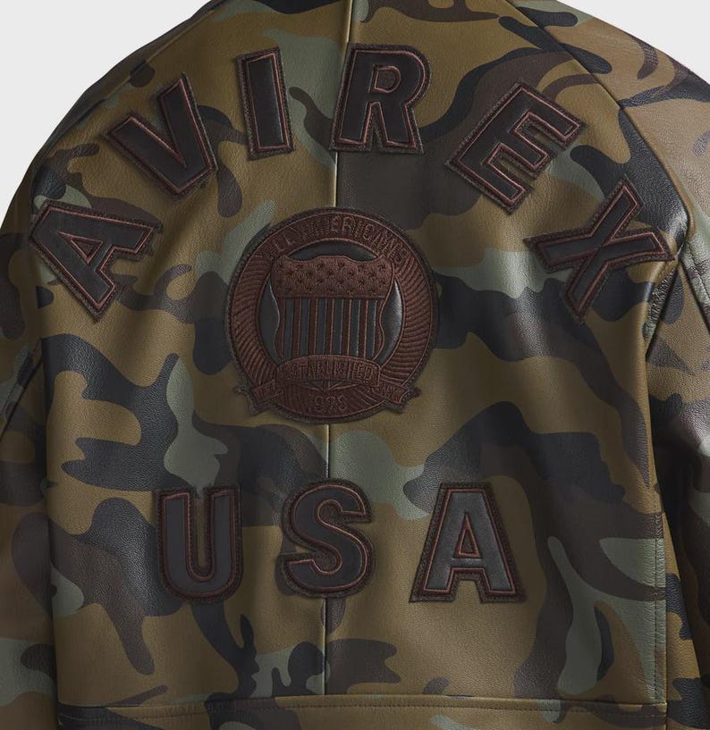 May include: A camouflage print jacket featuring dark brown embroidered lettering that spells AVIREX across the upper back and USA across the lower back. A circular brown patch in the center displays a shield emblem with the text ESTABLISHED 1975.