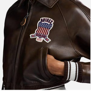 May include: A dark brown leather jacket featuring a front zipper closure and a ribbed hem with white stripes. An embroidered patch on the chest displays the text AVIREX, ESTABLISHED 1975, and a shield graphic with stars and red and white stripes.