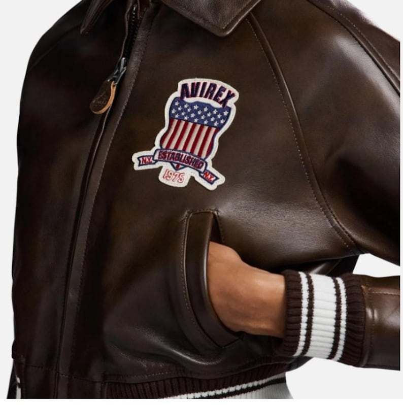 May include: A dark brown leather jacket featuring a front zipper closure and a ribbed hem with white stripes. An embroidered patch on the chest displays the text AVIREX, ESTABLISHED 1975, and a shield graphic with stars and red and white stripes.