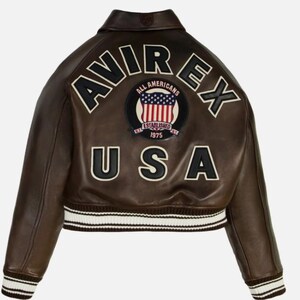 May include: A dark brown leather bomber jacket featuring a ribbed collar, cuffs, and hem with white stripes. The back displays the text AVIREX in large black letters, the word USA, and a circular patch with an American flag and text ALL AMERICANS.