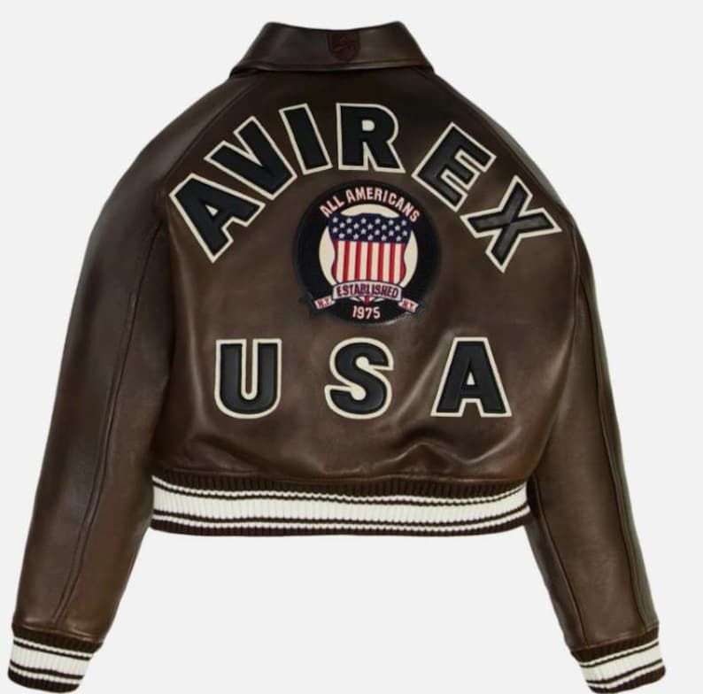 May include: A dark brown leather bomber jacket featuring a ribbed collar, cuffs, and hem with white stripes. The back displays the text AVIREX in large black letters, the word USA, and a circular patch with an American flag and text ALL AMERICANS.