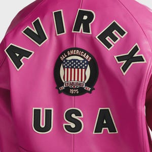 Op de afbeelding: Een felroze leren jas met zwart-witte geborduurde letters en patches. De jas heeft het woord "AVIREX" bovenaan, "USA" onderaan en een ronde patch met "ALL AMERICANS" en "ESTABLISHED 1975 N.Y."