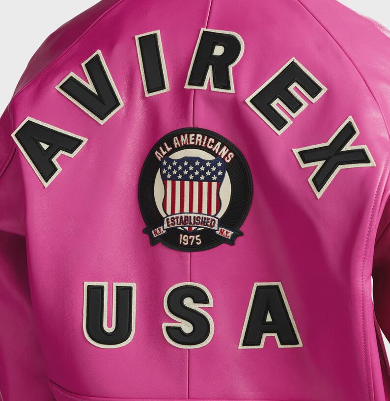 Op de afbeelding: Een felroze leren jas met zwart-witte geborduurde letters en patches. De jas heeft het woord "AVIREX" bovenaan, "USA" onderaan en een ronde patch met "ALL AMERICANS" en "ESTABLISHED 1975 N.Y."