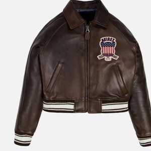 May include: A dark brown leather bomber jacket featuring a front zipper closure, a classic collar, and ribbed cuffs and hem with white stripes. An embroidered patch on the chest displays the text AVIREX above a stylized American flag design.